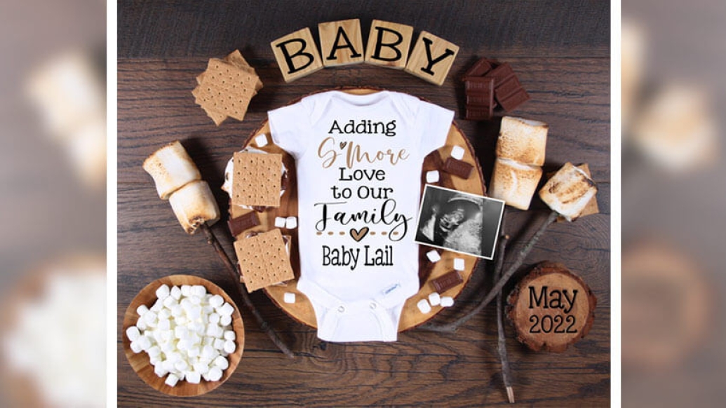 Baby onesie surrounded by smores, marshmellows and chocolate, reading adding smore love to our family baby Lail 