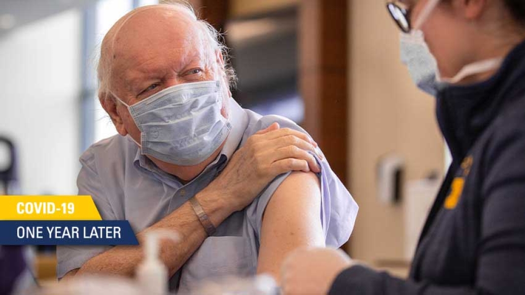 older man with white hair and blue face mask on lifting sleeve on blue shirt to get vaccine from worker